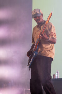 Male musician playing a blue bass guitar on stage amid pink and green lights.