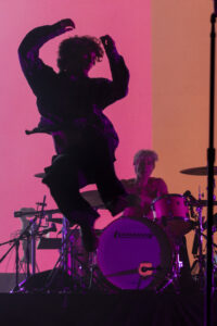 Person in a plaid shirt leaps mid-air on stage with a drummer and pink-orange backdrop behind them; dramatic silhouette and bright lighting.