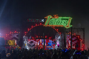 Neon festival sign reading 'The' in red script and large green letters followed by an orange arrow, glowily lit above a crowded nighttime stage.