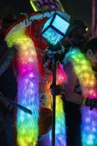 Person wearing a neon-lit feathered costume with a glowing cube headpiece at a night festival.