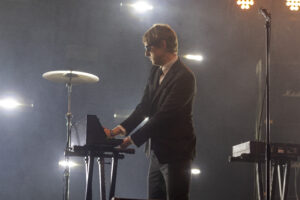 Male musician in a suit playing a keyboard on a smoky stage with drums in the background and bright lights overhead to suggest a live concert performance.