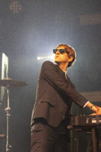 Male musician in a black suit wearing sunglasses, playing a keyboard on stage with bright backlight behind him.