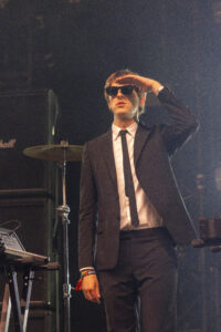 Male musician in a black suit and sunglasses saluting on stage with a drum kit behind him.