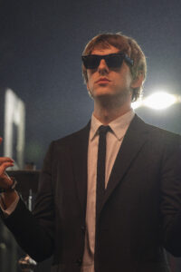 Man in a black suit, white shirt, and black tie wearing dark sunglasses on a dimly lit stage. He's positioned close to the camera with bright stage lights behind him, suggesting a performance setting.