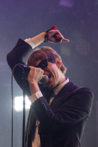 Male singer in a dark suit and sunglasses singing into a microphone with his right fist raised in the air on stage.