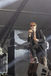 Male vocalist in a suit and sunglasses sings into a microphone on a concert stage with large speakers behind him.
