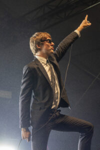 Male singer in a dark suit raises his right arm and microphone on stage in a concert setting.