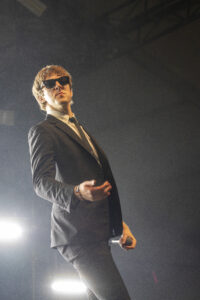 Male performer in a black suit and sunglasses on stage, holding a microphone, under bright stage lights.