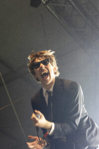 Male singer in a suit and sunglasses holding a microphone on stage, mid-performance with mouth open in song.
