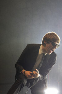 Male singer in a dark suit and sunglasses performing on stage, holding a microphone with bright lights behind him.