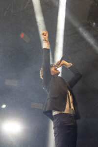 Male vocalist in a dark suit on stage, right fist raised as he sings into a microphone, with bright stage lights behind him.