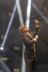 Male singer in a black suit and white shirt, wearing sunglasses, raising a microphone on a smoky concert stage with bright overhead lights