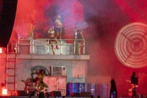 Performers dance on a two-level stage with red lighting and smoke, wearing vibrant outfits as a crowd watches below