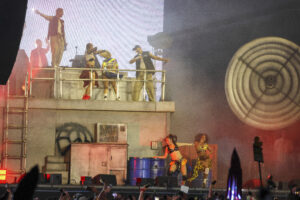 Live stage performance with dancers on a raised platform, crowd below, and a large circular prop on the right side.