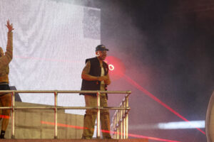 Male performer on a metal railing stage wearing a cap and vest, holding a microphone amid red lights and smoke.