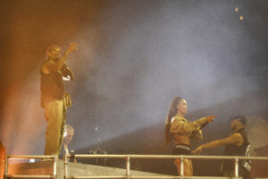 Male performer on the left raising his right hand and holding a microphone, bathed in orange stage light and fog, with a DJ in the background on a raised platform.