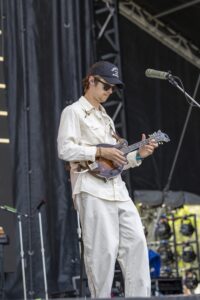 Musician wearing a cap and sunglasses, playing a mandolin on stage with a microphone nearby.