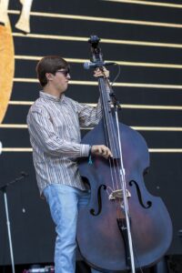 Musician wearing a striped shirt and sunglasses plays a double bass on a stage with a microphone nearby