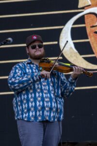 Musician wearing sunglasses and a patterned blue shirt plays a violin on stage with a microphone nearby.