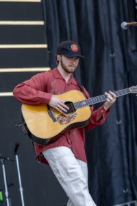 Male guitarist performing on stage in a red shirt and Tony Rice cap, playing an acoustic guitar.