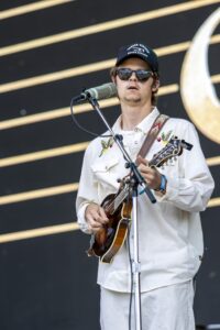 Male guitarist singing into a vintage-style microphone on stage, wearing sunglasses and a cap.