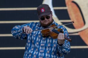 A bearded man in sunglasses and a patterned blue shirt plays a violin on stage with a dark backdrop behind him.
