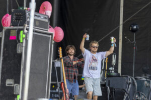 Two male musicians on a backstage stage, one raising a drink can, pink flamingo balloons and gear nearby.