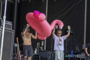 Two men on a backstage stage raise drinks beside a giant pink inflatable flamingo.