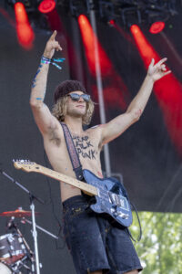 Shirtless guitarist on stage with sunglasses and a beanie, arms raised while playing a black electric guitar with blue paint on his arms.