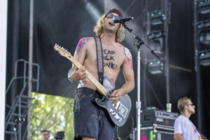 Shirtless guitarist wearing a knit cap and sunglasses while singing into a mic on stage, playing a black electric guitar with a decorated strap.