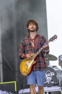 Male guitarist on stage playing a sunburst electric guitar, wearing a red plaid shirt and denim shorts at a live concert.