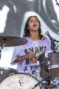 Female drummer singing into a microphone while playing a drum kit on stage, wet hair and a light purple shirt visible.