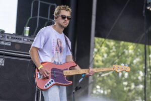 Male bassist on an outdoor stage, wearing sunglasses, playing a red Fender bass against a backdrop of amps and equipment.