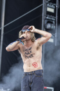 Male singer on a dark stage, shirtless and holding a microphone, wearing a knit cap and sunglasses on his head; text on chest reads 'CHEAP FOLK PUNK'.