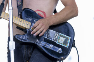 Guitarist playing a black electric guitar with stickers, shirtless chest with red paint circle, denim jeans in view.
