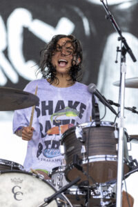 Female drummer with wet hair smiling as she plays a drum kit on stage, wearing a light purple shirt.