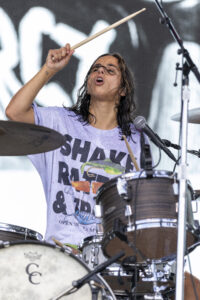 Female drummer mid-performance, raising a drumstick above her head while playing a kit on stage, with a microphone nearby and wet hair.
