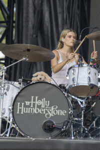 Female drummer with blonde braids in a white top playing a silver drum kit on stage, a teddy bear rests on the kit beside her.
