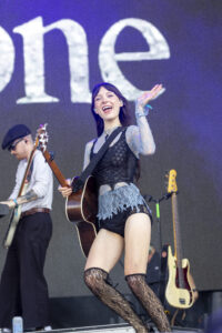 Woman guitarist performing on stage, smiling and waving, wearing a lace black top and thigh-high lace stockings, with a guitar strapped across her body.