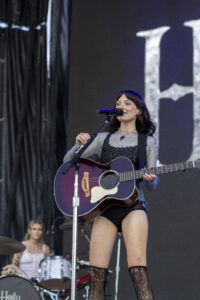 Female musician on stage singing into a microphone while playing a purple acoustic guitar with a guitar strap, wearing lace top and black shorts.
