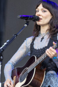 Female singer with brown hair performs on stage, singing into a microphone and playing an acoustic guitar.