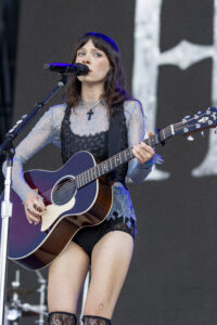 Female musician singing into a microphone while playing a guitar on stage, wearing a lace top and cross necklace.