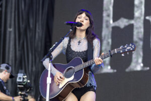 Female musician performing on stage with a purple acoustic guitar, singing into a microphone, wearing a blue lace dress and cross necklace.