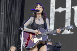 Female musician performing on stage, singing into a microphone while playing a red acoustic guitar with a cross necklace visible.