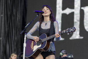 Female musician singing into a microphone while playing a purple acoustic guitar on stage.