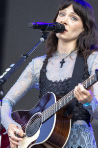 Singer performing on stage, speaking or singing into a microphone while playing an acoustic guitar.