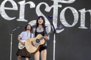 Smiling female musician playing an acoustic guitar on stage, with a large backdrop behind her.