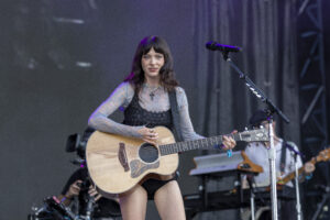 Female musician on stage playing an acoustic guitar, wearing a lace top and a cross necklace, with a microphone nearby.