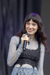 Female singer on stage holding a microphone, wearing a blue lace dress with a black corset, smiling as she sings into the microphone.