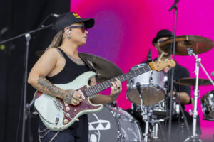 Female guitarist with tattoos, wearing a black cap, plays a mint-green Fender Stratocaster on stage with a bright pink backdrop behind her.